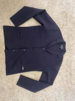 Svart kavaj från By Malene Birger - Stilren svart kavaj från By Malene Birger med tre knappar framtill och två fickor. Kavajen har klassisk krage och diskret logotypbroderi vid ena fickan. Materialet ser ut att vara stickat och mjukt, perfekt för en modern look.