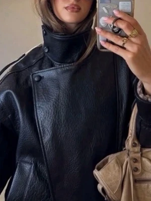 Svart oversized skinnjacka - Cool svart skinnjacka med hög krage och asymmetrisk knäppning. Jackan har en oversized passform, breda axlar och detaljer som knappar och fickor. Perfekt för dig som vill ha en edgy och trendig look.