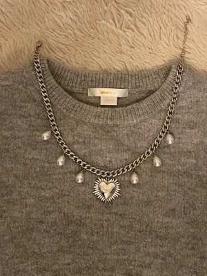 Silverfärgat halsband med pärlor och hjärta - Ett chunky silverfärgat rostfritt halsband med droppformade pärlor och ett stort hjärtformat hängsmycke med stråldetaljer. Kedjan är grov och har justerbar längd. Perfekt för att ge din outfit en cool och unik touch.