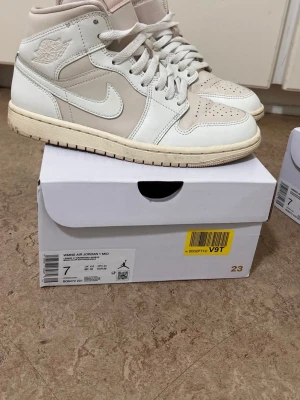 Air Jordan 1 Mid Beige/Vit - Säljer ett par Air Jordan 1 Mid sneakers i beige och vitt. Skorna har klassisk Jordan-design med läderpaneler, perforerad tå och ikonisk Swoosh på sidan. Mids höjd och bekväm dämpning. Perfekta för dig som vill ha en clean och stilren look.