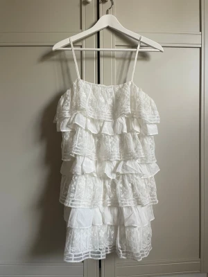 Vit volangklänning från Zara - Säljer en vit klänning från Zara med tunna axelband och flera lager av volanger. Klänningen har spetsdetaljer och en luftig, romantisk look som är perfekt för varma dagar. Materialet är bomull och polyester, vilket gör den både bekväm och lätt. 💗💗
