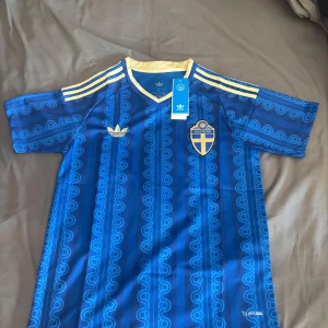 Blå Sverige bortställ tröja VM 2026  - Säljer en blå kortärmad Sverige tröja med gula detaljer och retroinspirerat mönster. Tröjan har gul krage, tre gula ränder på axlarna och Sveriges landslagsemblem på bröstet. Tillverkad i lätt och ventilerande material. 