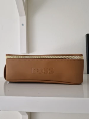 Brun necessär från BOSS Parfums - Snygg brun necessär från BOSS Parfums i slät konstläder med guldfärgad dragkedja. Rektangulär form med diskret BOSS-logga på sidan och präglad text på framsidan. Perfekt för att förvara småsaker eller smink i väskan. Aldrig använd. 