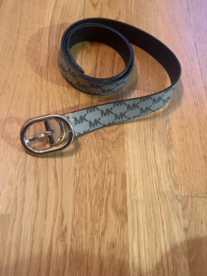 Beige MK bälte med logomönster (ny skick) - Snyggt beige bälte från Michael Kors med klassiskt MK-logomönster i brunt. Bältet har ett stilrent, rundat spänne i guld och är perfekt för att lyfta vilken outfit som helst. Passar både till jeans och kjol för en trendig look. 100% äkta nyskick.
