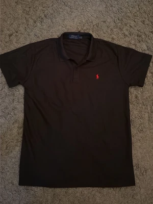 Svart polo t-shirt Ralph Lauren - Säljer en svart polo t-shirt från Ralph Lauren med  krage och korta ärmar. T-shirten har det röda Polo-loggan på bröstet och är tillverkad i bomull. Perfekt för den som gillar stilrena plagg med exklusiv känsla.