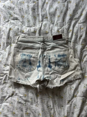 Ljusblå denimshorts med fransig kant - Säljer ett par ljusblå denimshorts från Hollister med coola blå tie-dye detaljer och fransig, rå kant nedtill. Klassisk femficksmodell med knapp och dragkedja. 