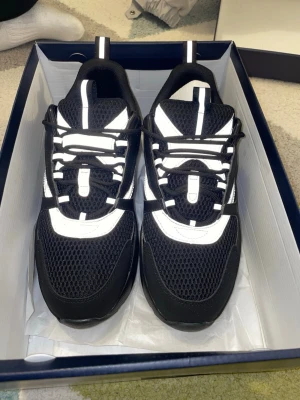 Svarta D**r b22 med grå detaljer - Snygga svarta sneakers med grå och vita detaljer. Ovandelen är i mesh och syntetmaterial för bra andningsförmåga och komfort. Skorna har en sportig look med reflekterande detaljer och klassisk snörning. Perfekta för dig som gillar stilrena och moderna sneakers.