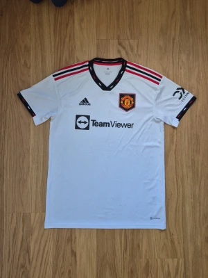 Manchester United retro bortatröja 22/23 Adidas - Snygg vit  retro Manchester United bortatröja från 22/23 sasöngen, Adidas med svarta och röda detaljer på axlarna. Tröjan har klubbmärke på bröstet, Adidas-logga och sponsortryck. Tillverkad i lätt och ventilerande material, perfekt för fotboll eller träning.