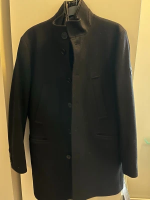 Svart ullkappa från J.Lindeberg - Stilren svart kappa från J.Lindeberg i ullmaterial med hög krage och knäppning framtill. Kappan har två snedställda fickor och en diskret logotyp på ärmen. Perfekt för dig som vill ha en klassisk och modern look. Kappan har en avtagbar väst på insidan 