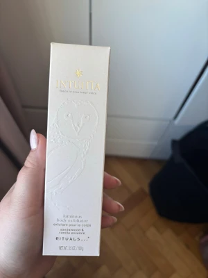 Rituals Intuita body exfoliator - Ljusbeige body exfoliator från Rituals Intuita med sandelträ och vaniljessens. Förpackningen har ett elegant ugglemotiv och guldtext. Perfekt för att ge huden en fräsch och mjuk känsla.