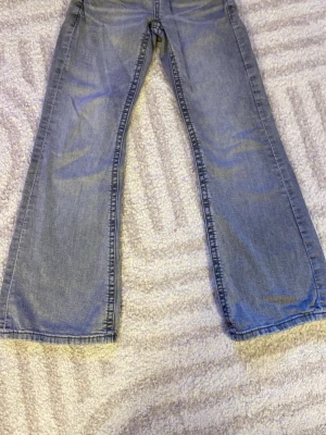 Ljusblå bootcut jeans - Säljer ett par ljusblå jeans med bootcut passform och klassiska fem fickor. Jeansen har en avslappnad look och är tillverkade i denim med synliga sömmar. Perfekta för dig som gillar en retro och bekväm stil. 