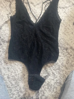 Svart spetsbody med djup urringning - Snygg svart body i spets med djup v-ringning och tunna axelband som korsas i ryggen. Bodyn har tryckknappar i grenen och ett blommigt spetsmönster framtill. Materialet är stretchigt och följsamt, perfekt för en trendig look.