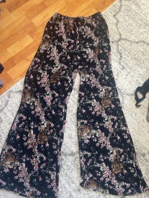 Svarta leggings med blommigt mönster - Säljer ett par svarta leggings med blommigt mönster och tigrar. De har vida ben och elastisk midja, perfekt för dig som gillar att sticka ut. Materialet känns mjukt och stretchigt, vilket gör dem riktigt bekväma. Passar dig som vill ha något coolt och unikt.