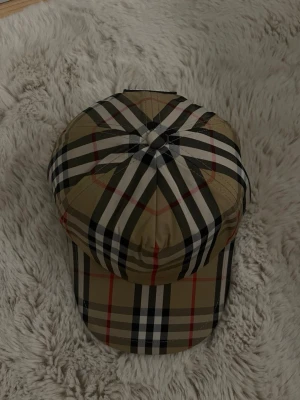 Beige keps med rutigt mönster Burberry - Snygg beige keps från Burberry med det klassiska rutiga mönstret i svart, vitt och rött. Kepsen har böjd skärm och justerbar passform. Perfekt accessoar för att lyfta din outfit med en lyxig touch.
