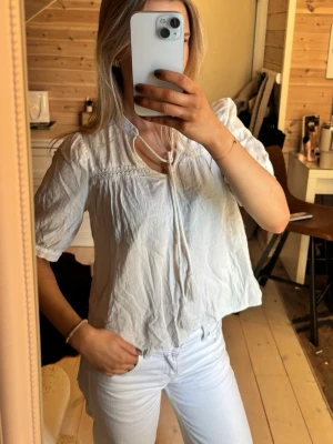 Vit boho-blus med knytband - Säljer en luftig vit blus i boho-stil med korta puffärmar och dekorativ spetsdetalj vid axlarna. Blusen har knytband vid halsen och är tillverkad i ett tunt, mjukt tyg som ger en avslappnad look. Perfekt att matcha med jeans eller kjol.