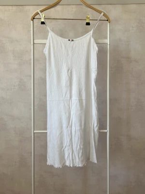 Vitt nattlinne med spets och rosett - Sött vitt nattlinne med tunna axelband och diskret spetskant upptill. Materialet har ett subtilt mönster och framtill sitter en liten rosa rosett med gröna blad. Lång och rak modell, perfekt för mysiga kvällar hemma.