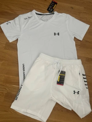 Vitt träningsset från Under Armour - Snyggt vitt träningsset från Under Armour med t-shirt och shorts. Båda plaggen har logga på bröst och ben samt tryck längs sidan. Tillverkade i lätt och snabbtorkande material, perfekt för gym eller löpning.