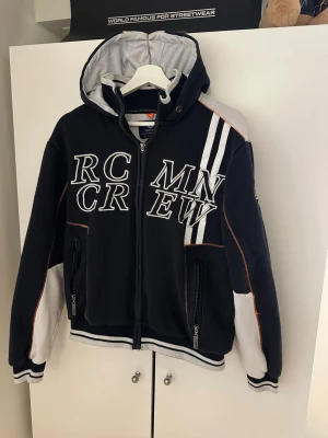 Marinblå zip hoodie med RC MN CREW-tryck - Snygg svart hoodie med vit och orange detaljsöm, stora broderade bokstäver 'RC MN CREW' framtill och dragkedja. Huva med vit insida och kontrastpaneler, samt randiga muddar. Två fickor med dragkedja och sportig streetwear-vibe.