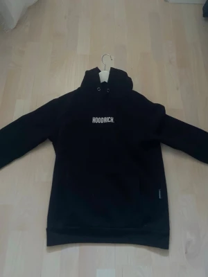 Svart hoodie från Hoodrich XS - Svart hoodie från Hoodrich med vit logotyp på bröstet. Tröjan har huva med snören och ribbade muddar vid ärmslut och nederkant. Perfekt för dig som gillar streetwear och vill ha en enkel men snygg look.