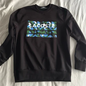 Svart Lacoste Tröja - En stilren svart sweatshirt från Lacoste med rund hals och ribbade muddar. På bröstet finns en färgstark grafisk logotyp i blå, gröna och vita nyanser som ger ett modernt och sportigt uttryck. Perfekt för en avslappnad men trendig vardagslook.