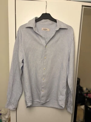 Ljusblå randig skjorta från Dressmann - Klassisk ljusblå skjorta från Dressmann med tunna vita ränder. Skjortan har lång ärm, krage och knäppning framtill. Perfekt för en clean och stilren look. Tillverkad i ett lätt och bekvämt tyg.