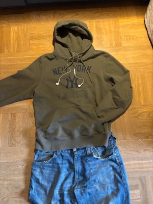 Olivgrön New York Yankees hoodie - Snygg olivgrön hoodie från New York Yankees med tryck framtill och klassisk känguruficka. Huva med dragsko och ribbade muddar. Perfekt för dig som gillar sportig streetstyle och vill ha något bekvämt och stilrent. Personen på bilden är 176 och väger 64 kg. Tröjan passar nog alla mellan 170-180 och 60-80kg!