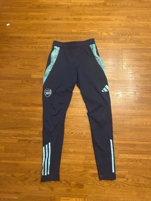 Arsenal träningsbyxor Adidas blå - Mörkblå träningsbyxor från Adidas med Arsenal-logga på vänster ben och ljusblå detaljer längs sidorna samt klassiska Adidas-ränder på benen. Byxorna har smal passform och är tillverkade i ett lätt och stretchigt material, perfekta för fotbollsträning.