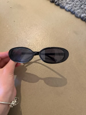 Svarta ovala solglasögon - Säljer ett par svarta solglasögon med oval form och stilren design. Glasen är mörka och bågarna är smala och minimalistiska, vilket ger en cool och modern look. Perfekta för att lyfta din outfit under soliga dagar.