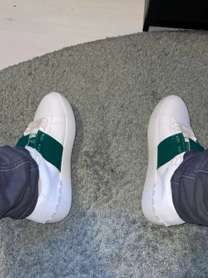 Valentino vita sneakers med grön detalj - Snygga vita sneakers från Valentino med en bred grön panel över snörningen och klassisk logga på tungan. Skorna har en låg profil och är tillverkade i slätt läder med vita skosnören. Perfekt för dig som vill sticka ut med stilrena detaljer.