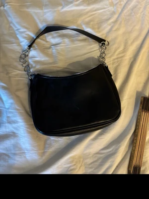 Svart axelremsväska med kedjedetaljer - Snygg svart handväska i slätt konstläder med silverfärgade kedjedetaljer på axelremmen. Väskan har en rundad form och dragkedja upptill. Perfekt för dig som gillar en stilren och trendig look. Använd 2 gånger. 