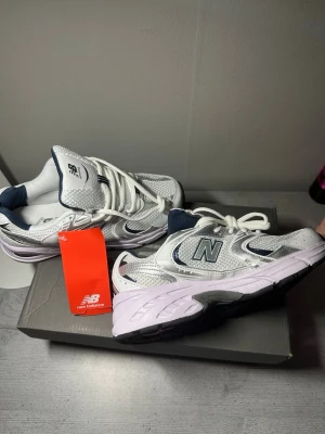 New Balance vita sneakers - Snygga vita New Balance sneakers med mesh och läderdetaljer, mörkblå inslag och silverfärgade accenter. Klassisk låg modell med ABZORB-sula för extra komfort. Perfekta för dig som gillar sportig och trendig stil.