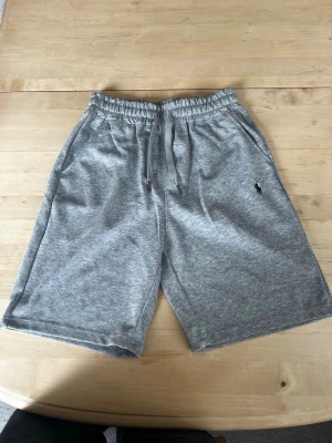 Grå shorts från Polo Ralph Lauren - Snygga grå shorts från Polo Ralph Lauren med klassisk broderad logga på benet. De har elastisk midja med snörning och är tillverkade i mjuk bomull, perfekta för en avslappnad stil. Två sidofickor och en bekväm passform.