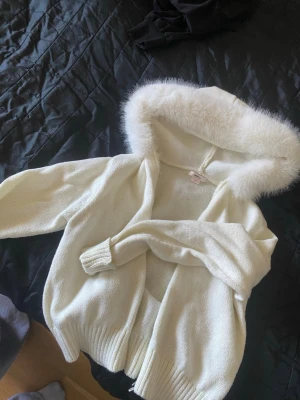 Vit kofta med fluffig huva - Snygg vit kofta med långärmad design och en stor huva med fluffig fuskpäls. Koftan är stickad och har ribbade muddar vid ärmslut och nederkant. Perfekt för att hålla sig varm och se stylish ut under kyliga dagar. 