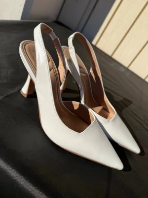 Vita slingback pumps med klack från k.cobler - Säljer ett par stilrena vita pumps med spetsig tå och öppen häl med slingback-rem från k.cobler. Otroligt sköna, har samma i svart och kan gå hela kvällar utan några problem. Perfekta till studenten, midsommar och sommaren🌸