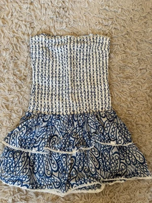 Blåvit klänning med volanger - Säljer en söt blåvit klänning med elastisk smock upptill och två lager volanger nedtill. Toppen har ett unikt mönster i blått på vit botten och spetsdetaljer längs kanterna. Perfekt för sommarens alla tillfällen!