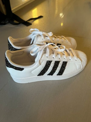 Adidas Superstar vita sneakers med glitter - Säljer ett par vita Adidas Superstar sneakers med svarta glitterdetaljer på de klassiska tre ränderna och hälen. Skorna har vit gummisula, tåhätta i snäckform och vita skosnören. Perfekta för dig som vill ha en sportig look med extra bling. Storlek 39, fotlängd 24 cm, aldrig använt. De är handgjorda därför dyrare 