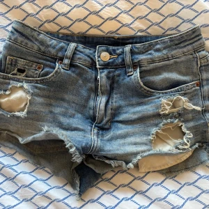 Blå slitna jeansshorts med rå kant - Säljer ett par blå jeansshorts med rå kant och slitna detaljer på framsidan. Klassisk femficksmodell med knappstängning och ljus tvätt. Perfekta för en avslappnad och trendig look under varma dagar.