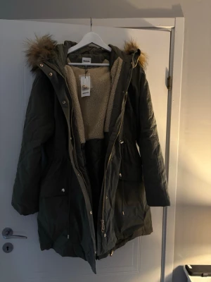 Olivgrön parkajacka från Indiska - Parkajacka i olivgrönt från Indiska med mysigt beige teddyfoder och huva med pälsdetalj. Jackan har dragkedja, knappar och stora fickor. Perfekt för kalla dagar och har en avslappnad passform. Helt oanvänd, med alla lappar kvar.