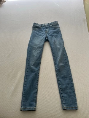 Blå skinny jeans med klassisk passform - Säljer ett par blå skinny jeans med klassisk femficksdesign och raka ben. Jeansen har knapp- och dragkedjeförslutning samt detaljerade sömmar. Materialet är denim och färgen är ljusblå. Perfekt för dig som gillar en enkel och stilren look.
