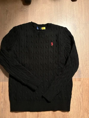 Svart kabelstickad tröja Polo Ralph Lauren - Svart kabelstickad tröja från Polo Ralph Lauren med klassisk rund halsringning och röd broderad logotyp på bröstet. Tröjan har långa ärmar och ribbade muddar. Perfekt för dig som gillar stilrena och tidlösa plagg.