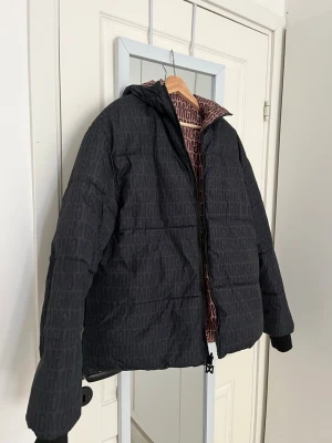 Svart pufferjacka från Missguided - Säljer en svart pufferjacka från Missguided med diskret logomönster över hela jackan. Jackan har huva, dragkedja framtill och ribbade muddar vid ärmsluten. Vändbar jacka/modell.