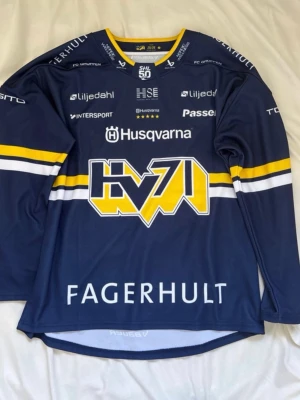 HV71 matchtröja - HV71 hockeytröja i blått med gula och vita ränder, tillverkad av funktionsmaterial. Stort HV71-tryck på bröstet, flera sponsorloggor och lång ärm. Perfekt för dig som älskar hockey och vill visa vilket lag du hejar på.