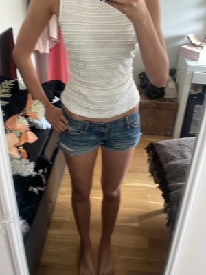 Vit ribbad linnetopp med knytband - Supersnygg vit ribbad linnetopp med hög hals och breda axelband. Toppen är ärmlös och har ett brett knytband i ryggen som ger en öppen rygg och en unik look. Perfekt till sommaren och lätt att matcha med jeansshorts eller kjol.