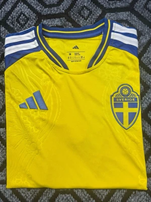 Svenska VM tröjan hemma - Hemma svensk landslagströja med blå detaljer och vita ränder på axlarna. Broderad Sverige-logga på bröstet och Adidas-logga i blått. Tillverkad i lätt och ventilerande material, perfekt för fotboll eller träning.