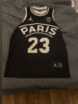 Jordan x PSG basketlinne svart - Snyggt svart basketlinne från Jordan x PSG med vita detaljer. Framsidan har stor text 'PARIS' och siffran 23, samt Jordan-logga och PSG-märke. Baksidan har 'JORDAN' och siffran 23. Perfekt för basket eller streetwear.