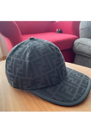 Svart keps från Fendi med FF-mönster - Säljer en svart keps från Fendi med det klassiska FF-logomönstret över hela kepsen. Kepsen har böjd skärm med en diskret kant i svart och är tillverkad i ett tygmaterial som ger en lyxig känsla. Perfekt för dig som vill ha en stilren och exklusiv accessoar.