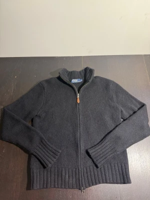Mörkgrå stickad ziptröja från Polo - Svart stickad tröja från Polo Ralph Lauren med hel dragkedja och hög krage. Tröjan har ribbade muddar vid ärmslut och nederkant samt en stilren, minimalistisk look. Perfekt för lager-på-lager och enkel att matcha.