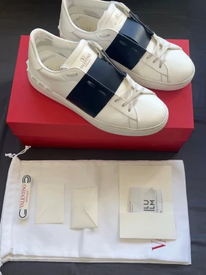 Valentinos open mörkblå - Snygga vita sneakers från Valentino med en bred svart läderdetalj över vristen. Klassisk låg modell med rund tå och vita snören. Märkeslogga på plösen och insidan. Kommer med originalkartong och dustbag.