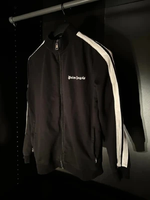 Svart track jacket från Palm Angels - Svart track jacket från Palm Angels med vita ränder längs ärmarna och dragkedja framtill. Jackan har hög krage, ribbade muddar och logga på bröstet. Perfekt för en sportig och trendig streetwear-look.