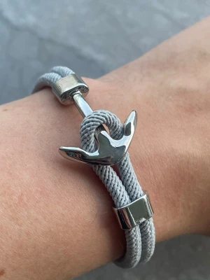 Grått armband med ankardetalj - Snyggt armband i grått rep med en blank silverfärgad ankare som lås. Armbandet har dubbla rep och metalliska detaljer som ger en modern och stilren look. Perfekt accessoar för dig som gillar marina vibbar och enkel design.